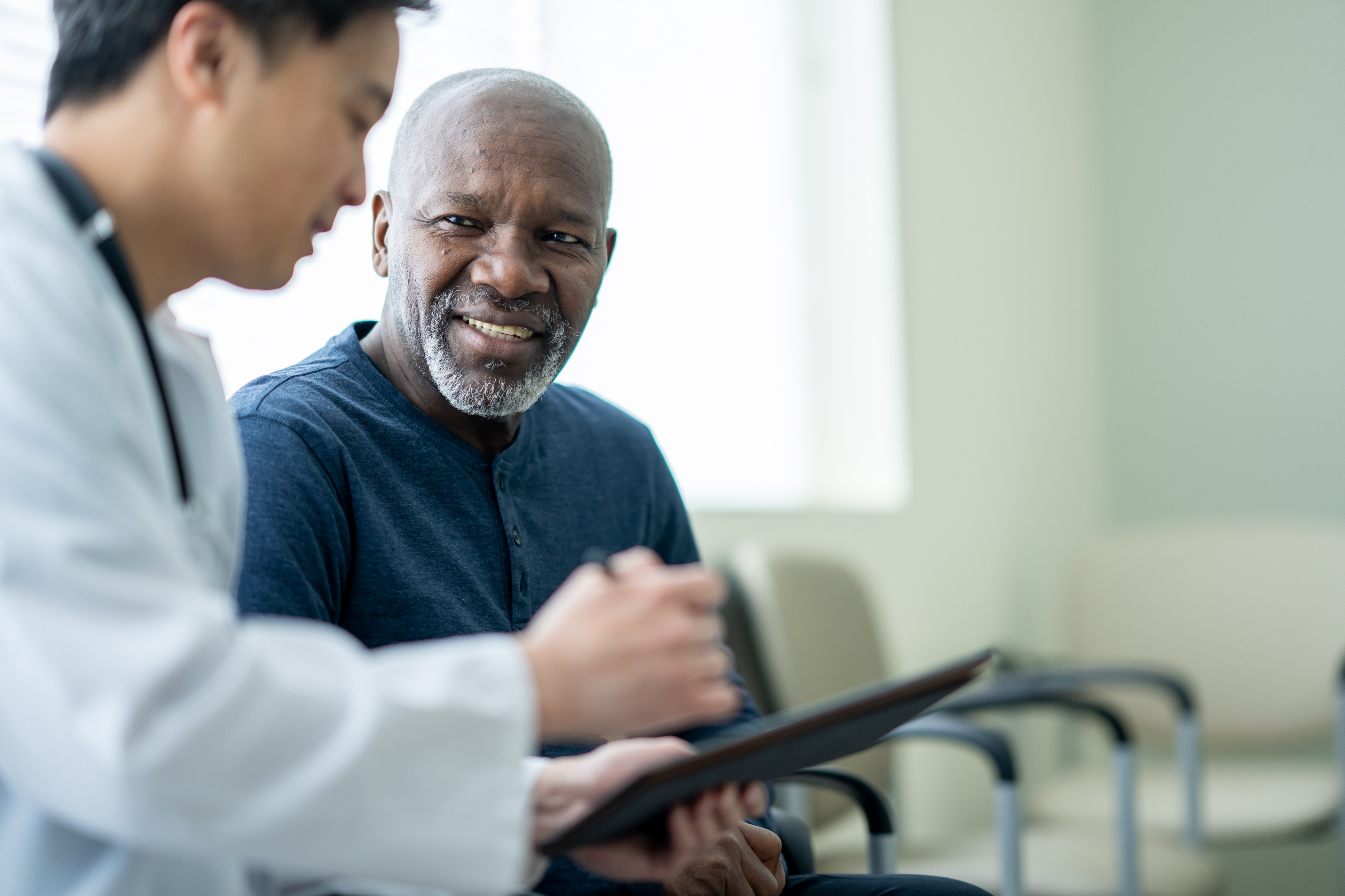 A senior gentleman sits in a waiting room beside his doctor